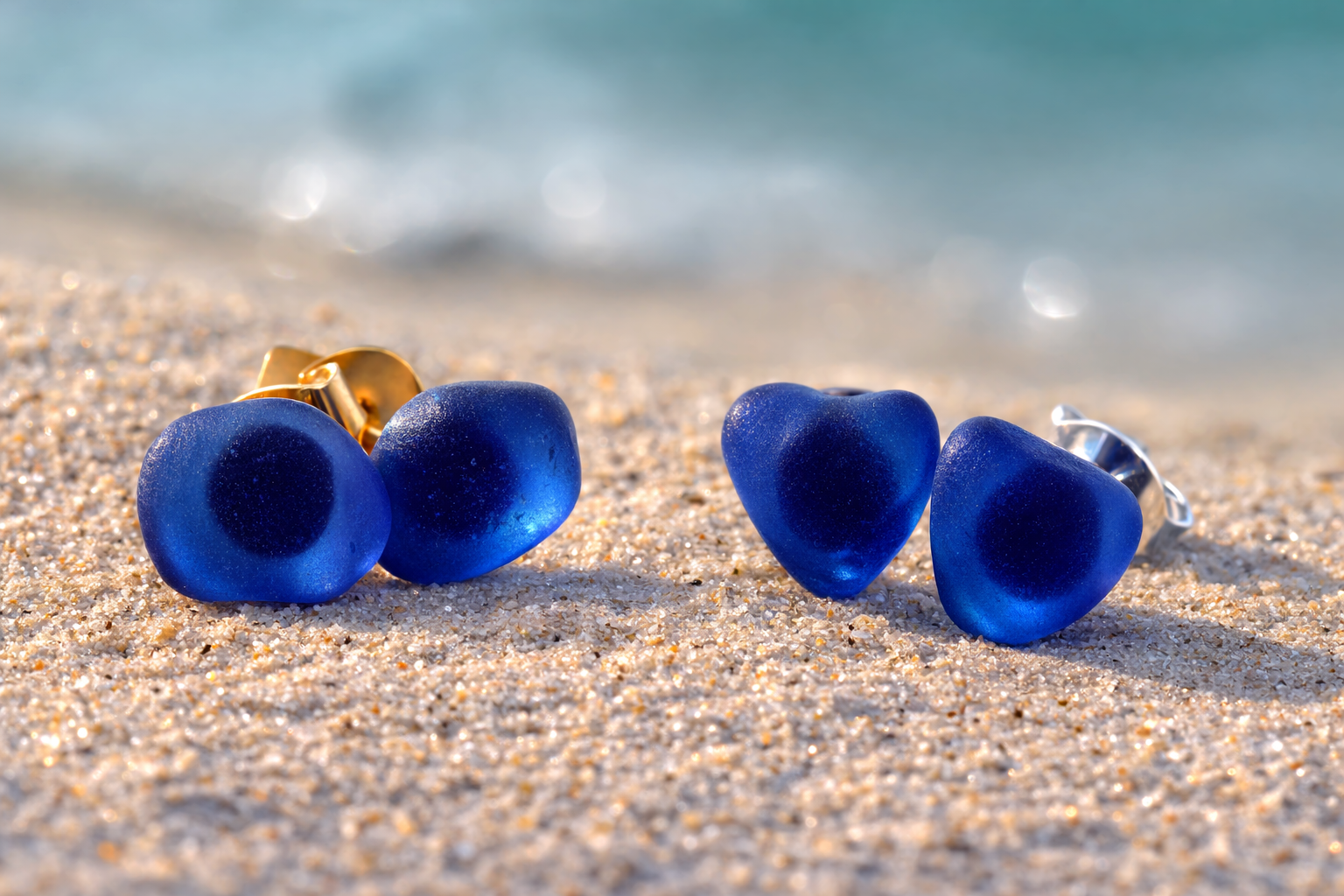 Blue heart-shaped earrings on a sandy beach with ocean in the background