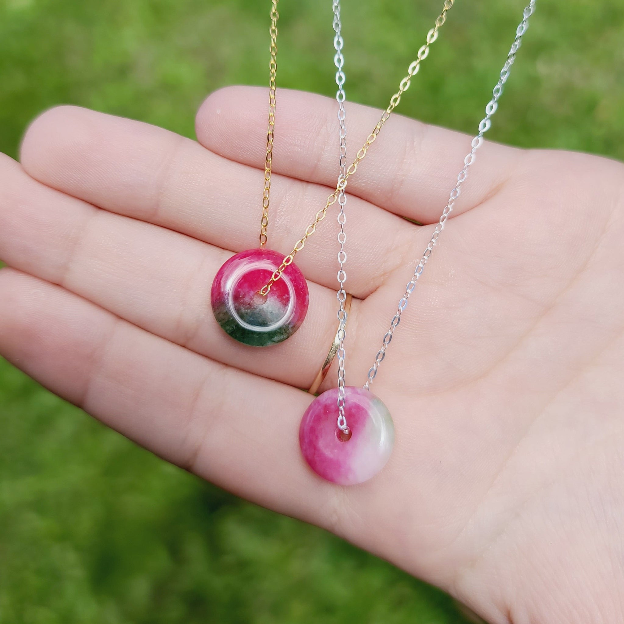 Two Jade necklaces with red and green coloring, held in a person's hand, with gold-filled or sterling silver chains.